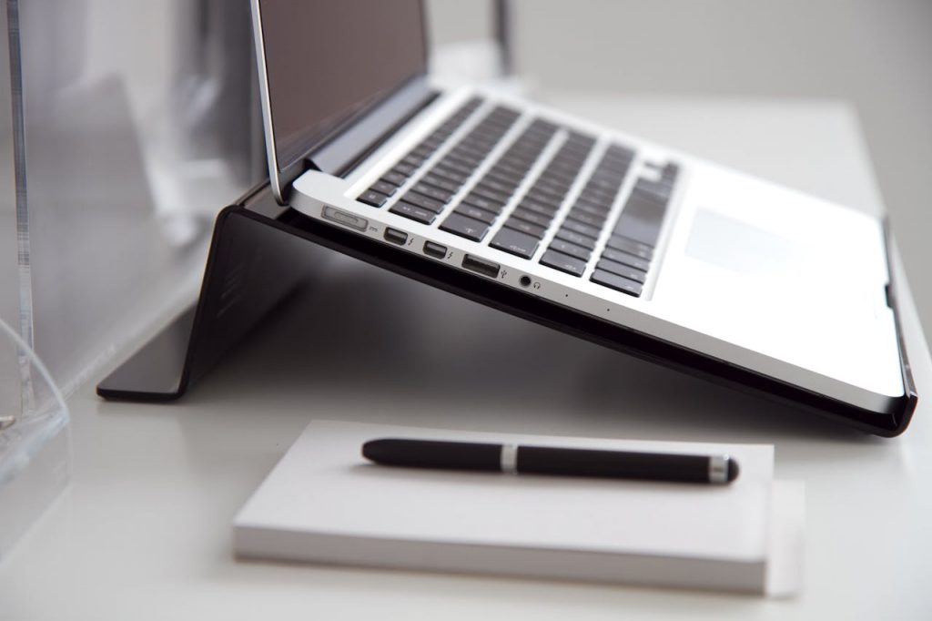 The Art of Drawing Readers In: Your attractive post title goes here Side view of a laptop on a stand with a notebook and pen on a white desk.